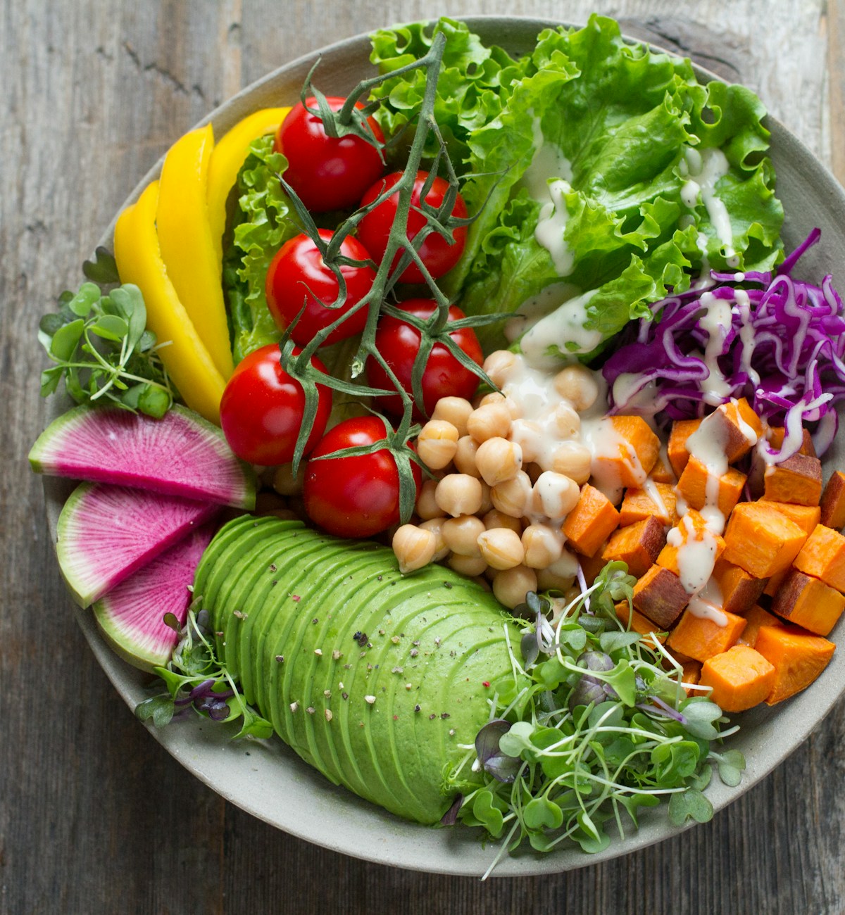 Overhead view of a raw vegetable salad bowl with chickpeas, avocado, cherry tomatoes, lettuce, sweet potato, and purple cabbage on a weathered wood board